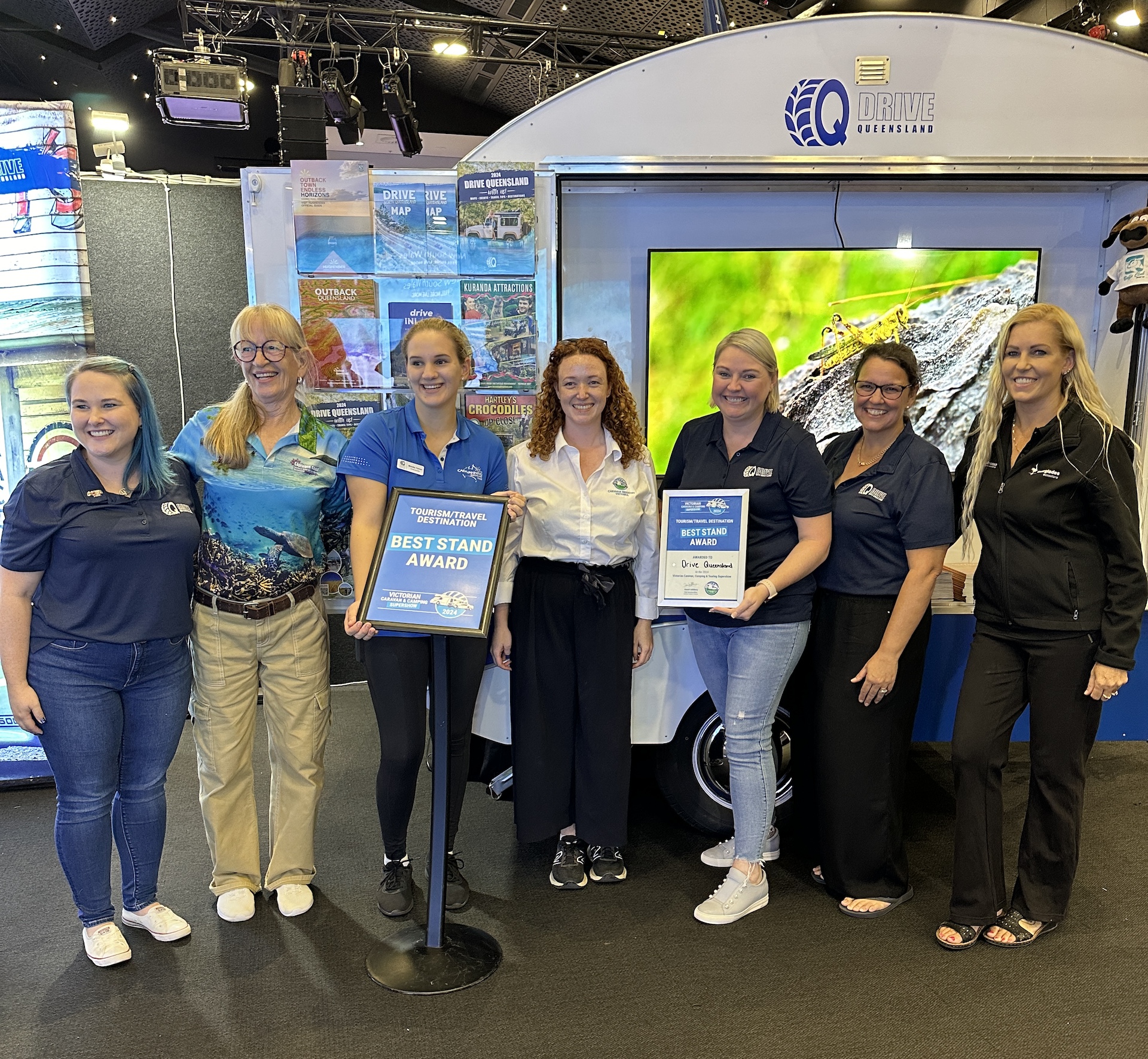 A group of seven individuals stands indoors at an event, posing with an award and a certificate in front of a display booth.