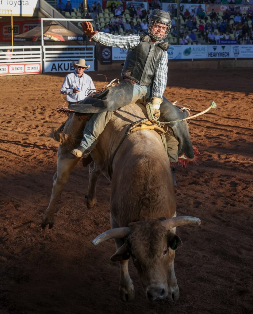 A rodeo rider in safety gear balances on a bucking bull in a dirt arena as a rodeo clown stands nearby, with spectators watching from the stands.