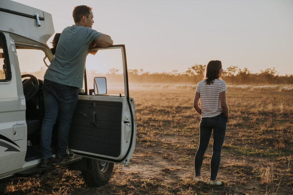 A man leans on a car door, while a woman stands nearby facing an open field at sunset.