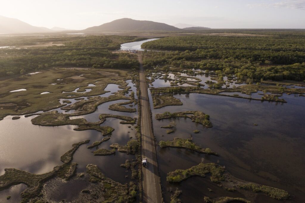 Aerial view of a road cutting through wetlands, with a river and mountains in the background under a clear sky.