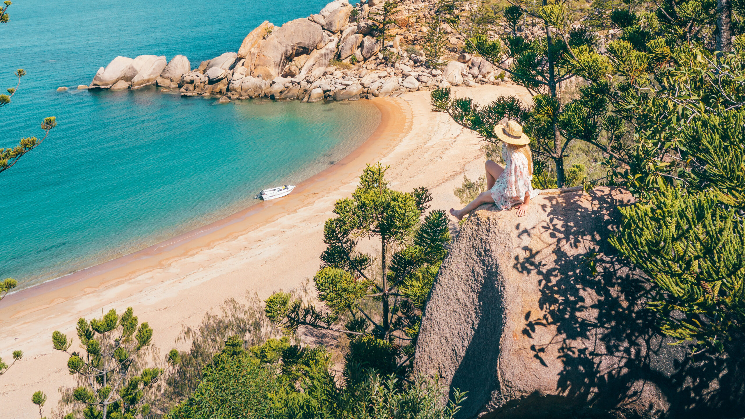 Person sitting on a rock overlooking a tranquil beach with turquoise water, surrounded by greenery and rocks. A small boat is on the sand near the shoreline.