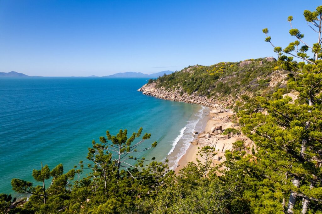 Coastal view of a rocky beach with turquoise water, surrounded by green trees under a clear blue sky.