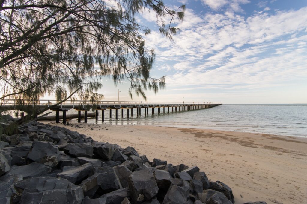 A long pier extends over the water from a sandy, rocky beach, with a tree in the foreground and a partly cloudy sky above.