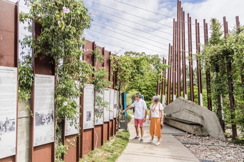 Two people walk through an outdoor exhibit with informational panels, tall rust-colored structures, and greenery on a sunny day.