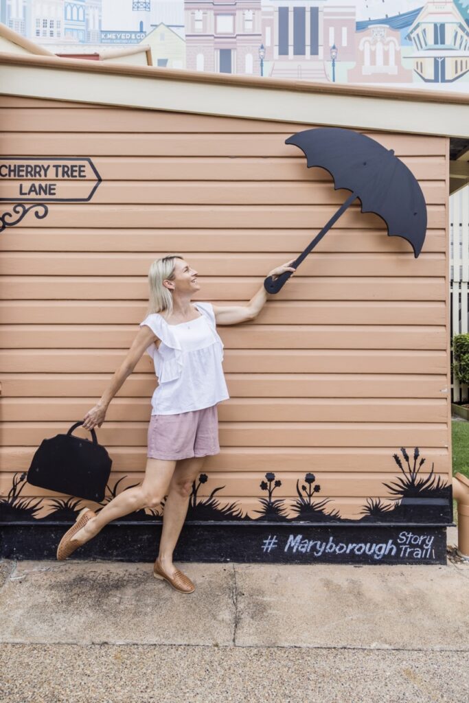 A woman in a white blouse and pink shorts poses playfully, pretending to hold onto a painted black umbrella on a wall mural in front of a sign reading 