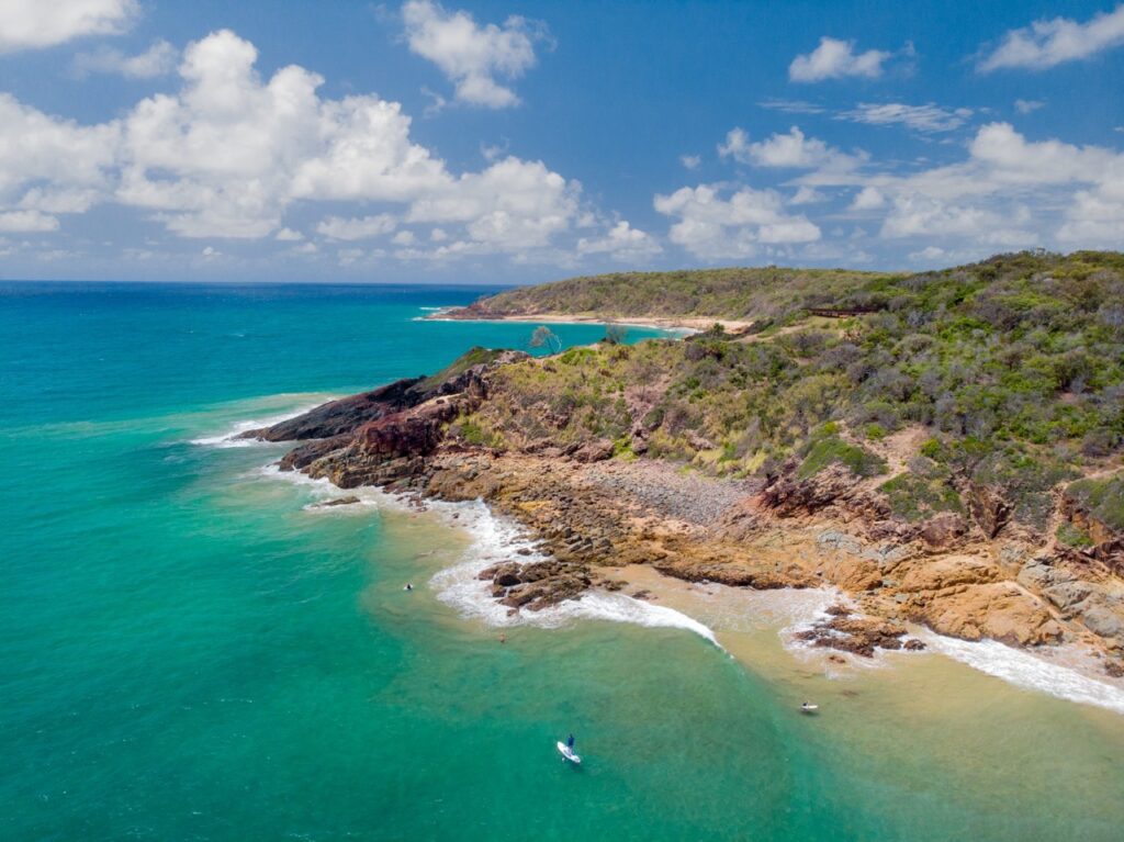 Aerial view of a coastal landscape featuring rocky shores, lush greenery, and turquoise ocean waters under a partly cloudy sky.