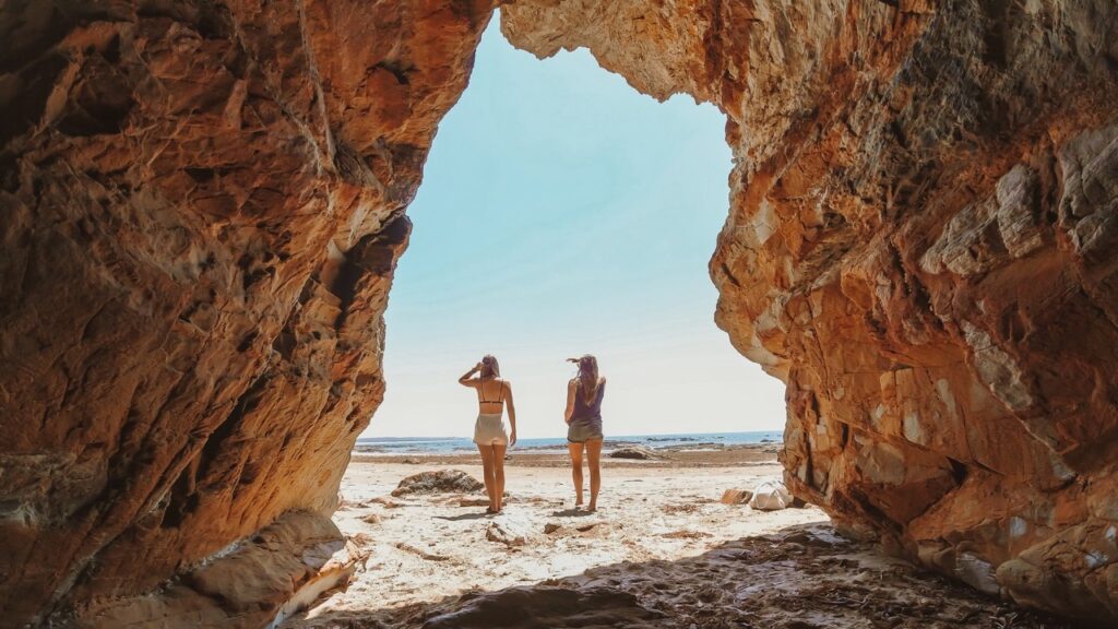 Two people in swimwear stand on a sandy beach, viewed from inside a rocky cave opening with a clear sky and ocean in the background.