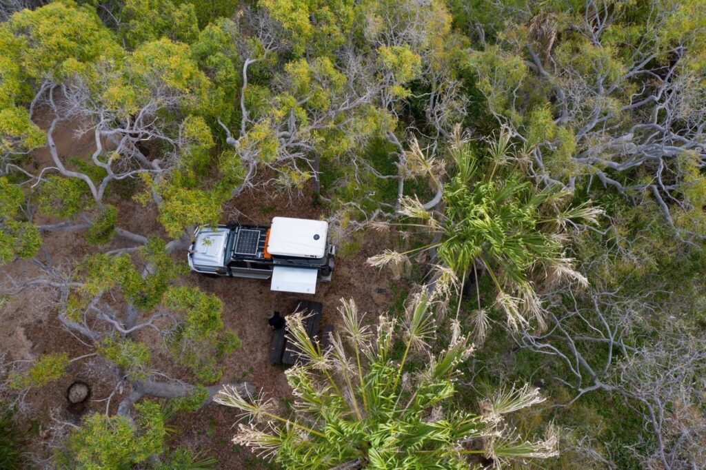 Aerial view of a white off-road vehicle with rooftop equipment parked among dense, green trees in a forested area.