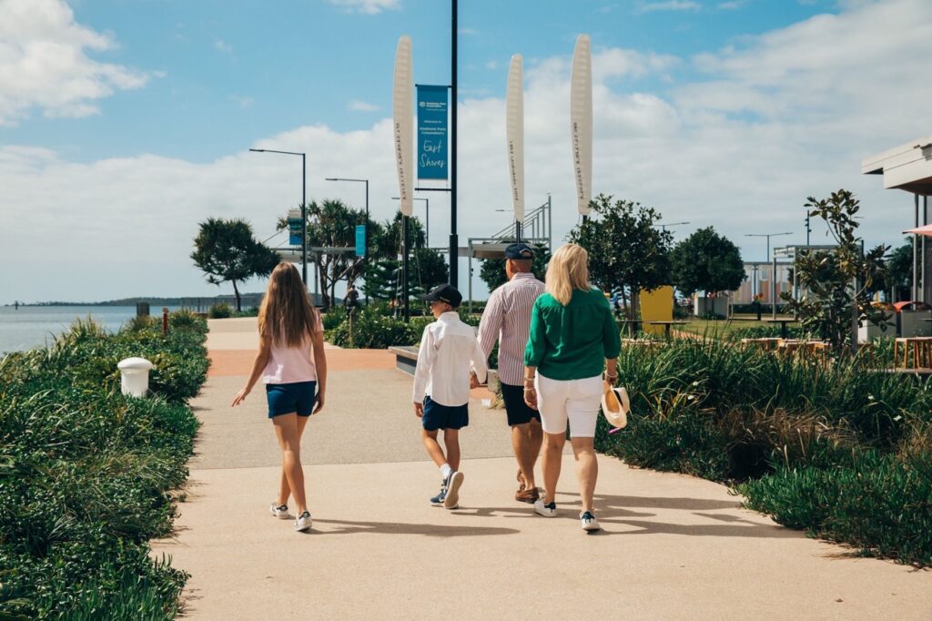 A group of four people, including two adults and two children, walk together along a sunny, landscaped waterfront pathway.