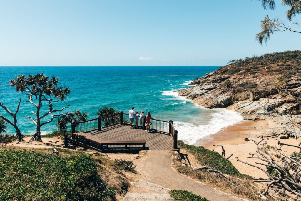 Three people stand on a wooden lookout platform overlooking a coastal scene with waves crashing onto a rocky shore. The sky is clear and the ocean is blue.