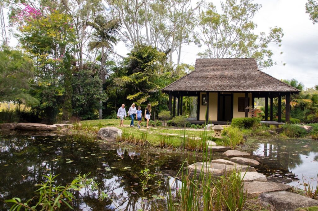 A family walks along a stone path near a pond, approaching a rustic pavilion surrounded by lush greenery and tall trees.
