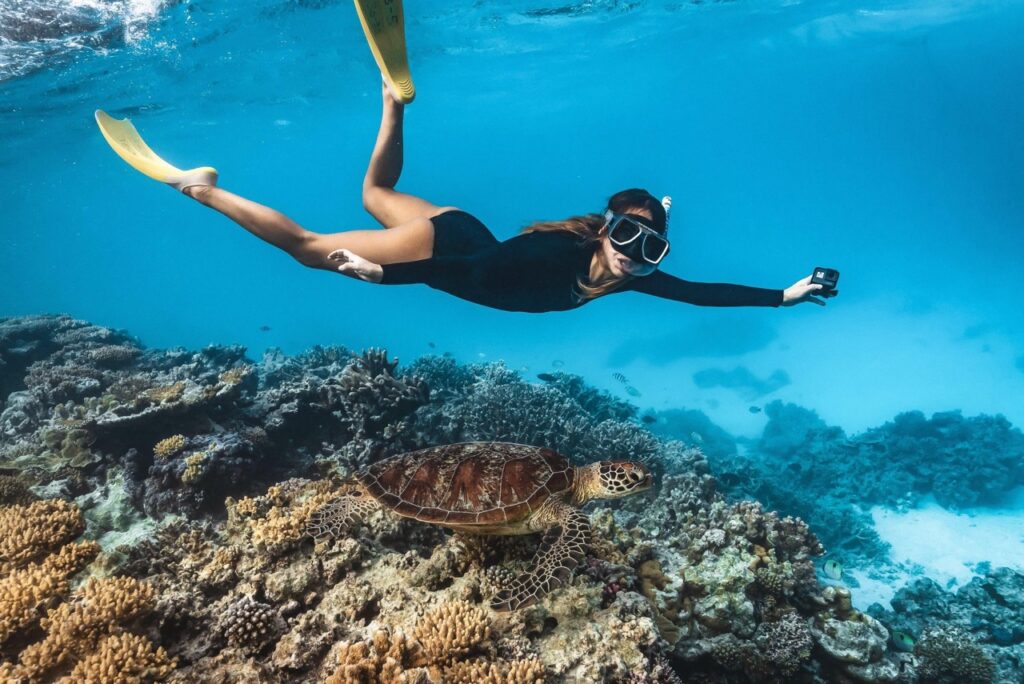A person in a black wetsuit and yellow flippers swims underwater over a coral reef, holding a camera and observing a sea turtle below.