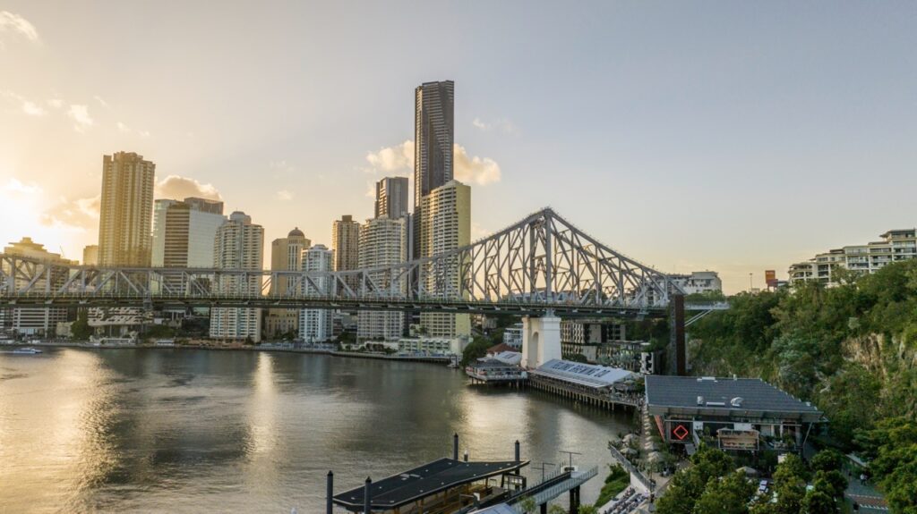 A bridge spans a river in front of a city skyline with tall buildings at sunset, with both greenery and urban structures visible in the foreground.