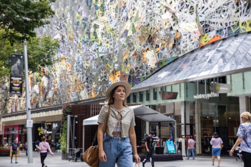 A person in casual attire and a hat walks through a busy outdoor shopping area with colorful, intricate wall art in the background.