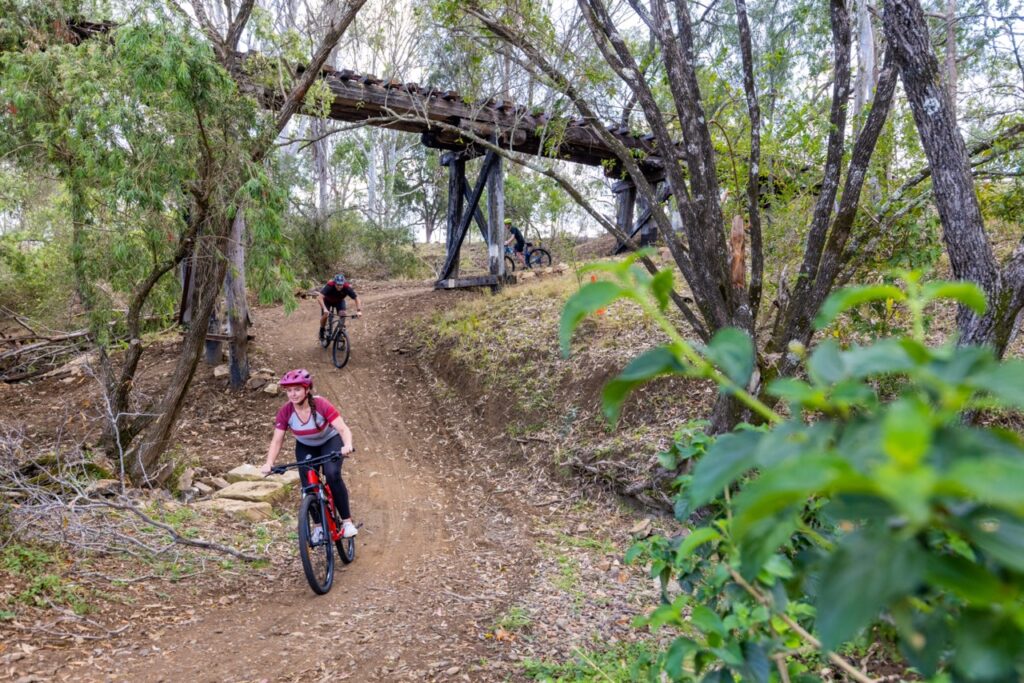 Three people ride bicycles on a dirt trail under an old wooden bridge surrounded by trees. One person leads, wearing a red helmet and black outfit, while the others follow at a distance.