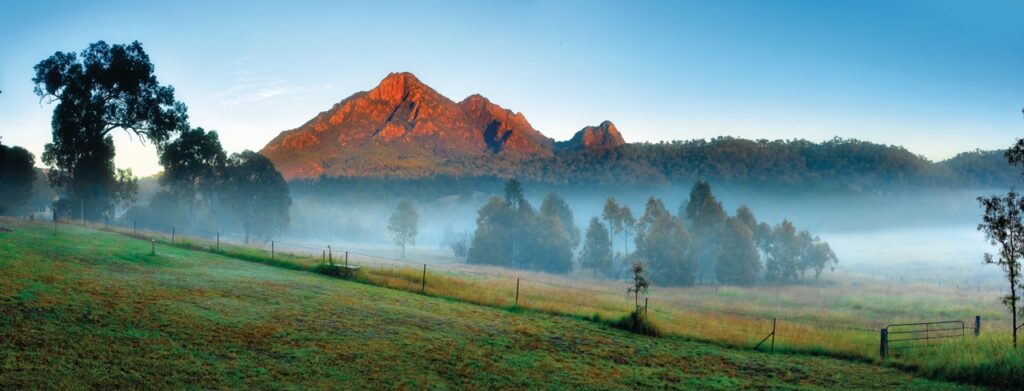 A mountain view at sunrise with the peak illuminated, surrounded by mist and trees in the foreground, and a fence in the lower right corner.