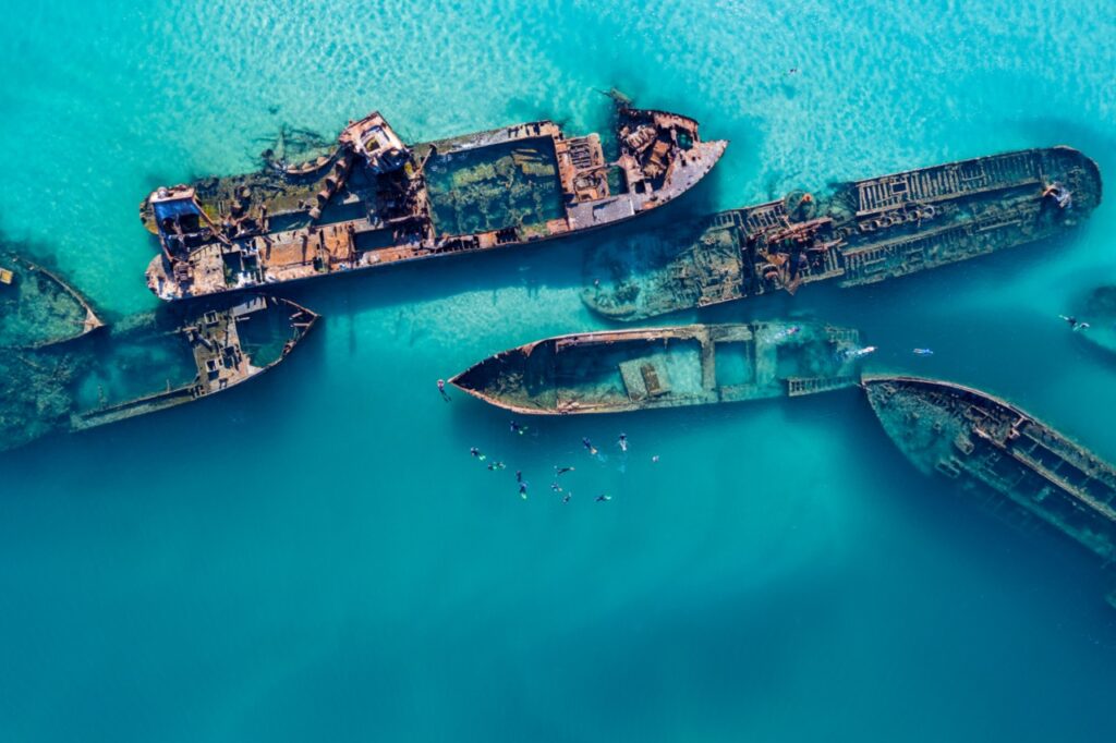 Aerial view of sunken ships submerged in clear turquoise water, with small groups of people swimming around the wreckage.