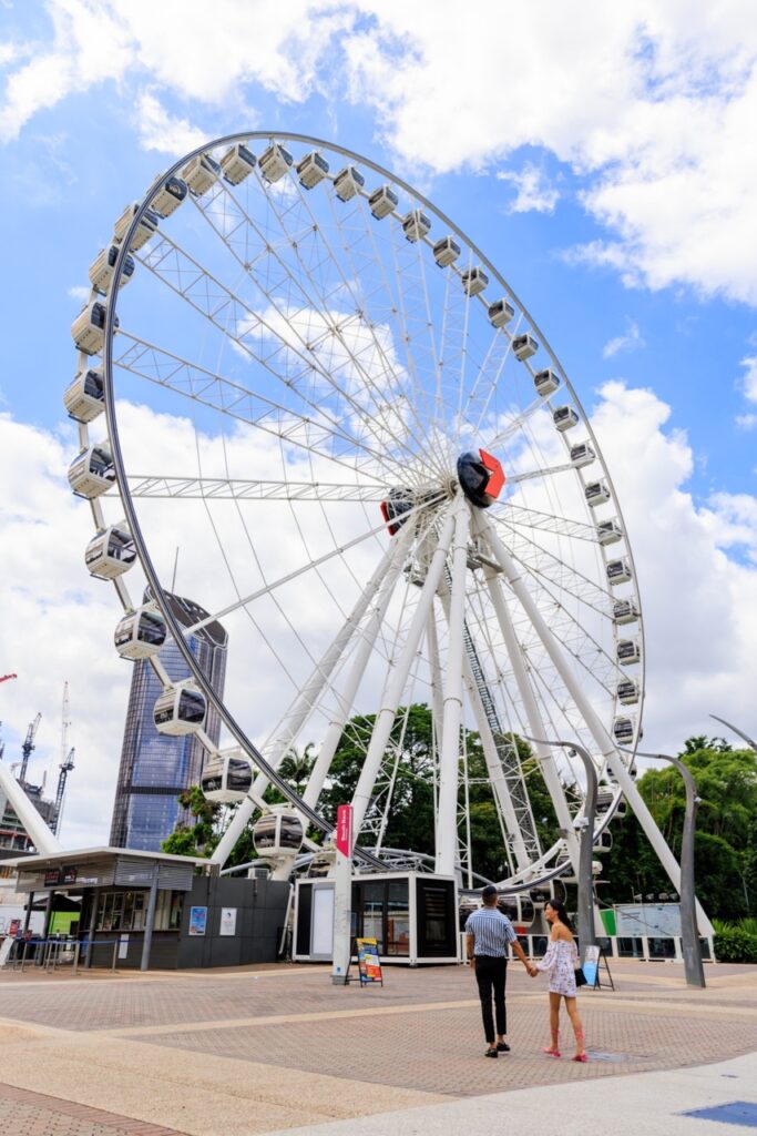 A large Ferris wheel stands against a blue sky with scattered clouds. Two people walk towards the entrance area below.