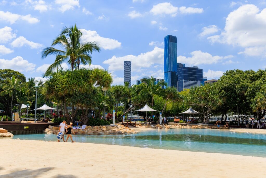 A beach and lagoon with palm trees in the foreground and a city skyline with tall buildings in the background under a blue sky with clouds.