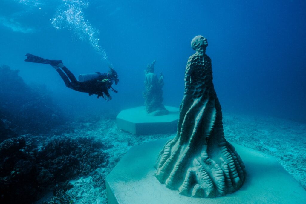 A scuba diver explores an underwater art installation featuring human-like statues on pedestals in a blue ocean setting.