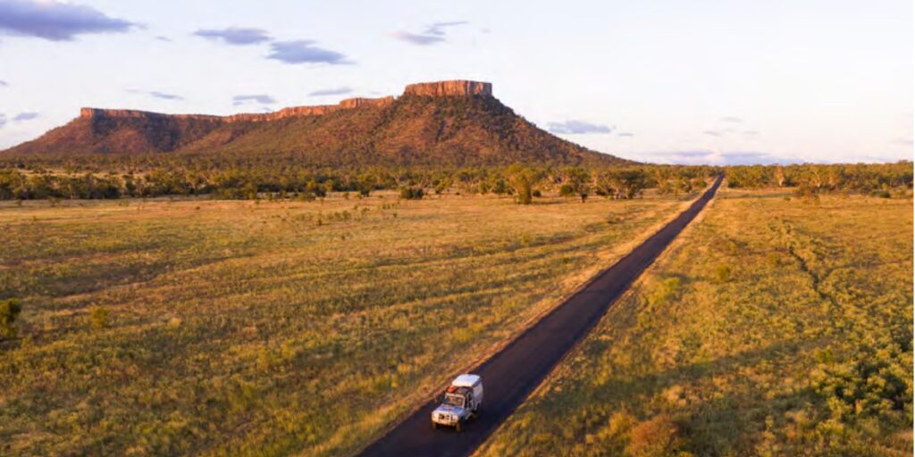 A vehicle travels on a long road through open grassland with a plateau in the background under a clear sky.