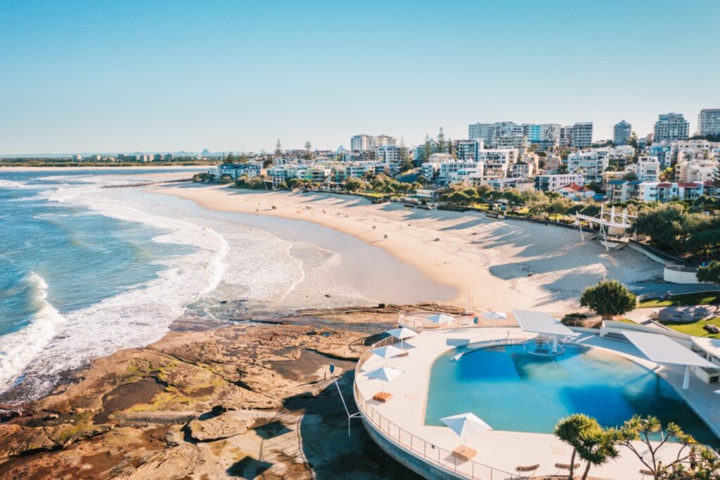 A coastal cityscape features a beachfront pool with white shade structures, waves crashing on a sandy beach, and modern buildings in the background under a clear blue sky.