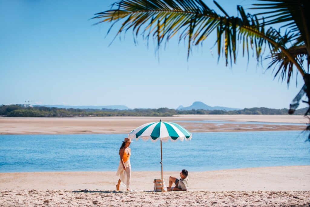 Two people on a sandy beach. One is standing, and another is seated under a green and white umbrella with a water body and mountains in the background, framed by a palm tree.