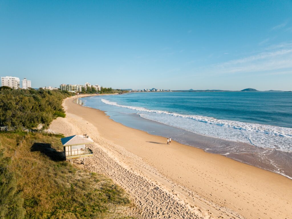 Aerial view of a sandy beach with a small white structure in the foreground and a cityscape in the background. The ocean waves gently reach the shore, and two people are walking near the water.