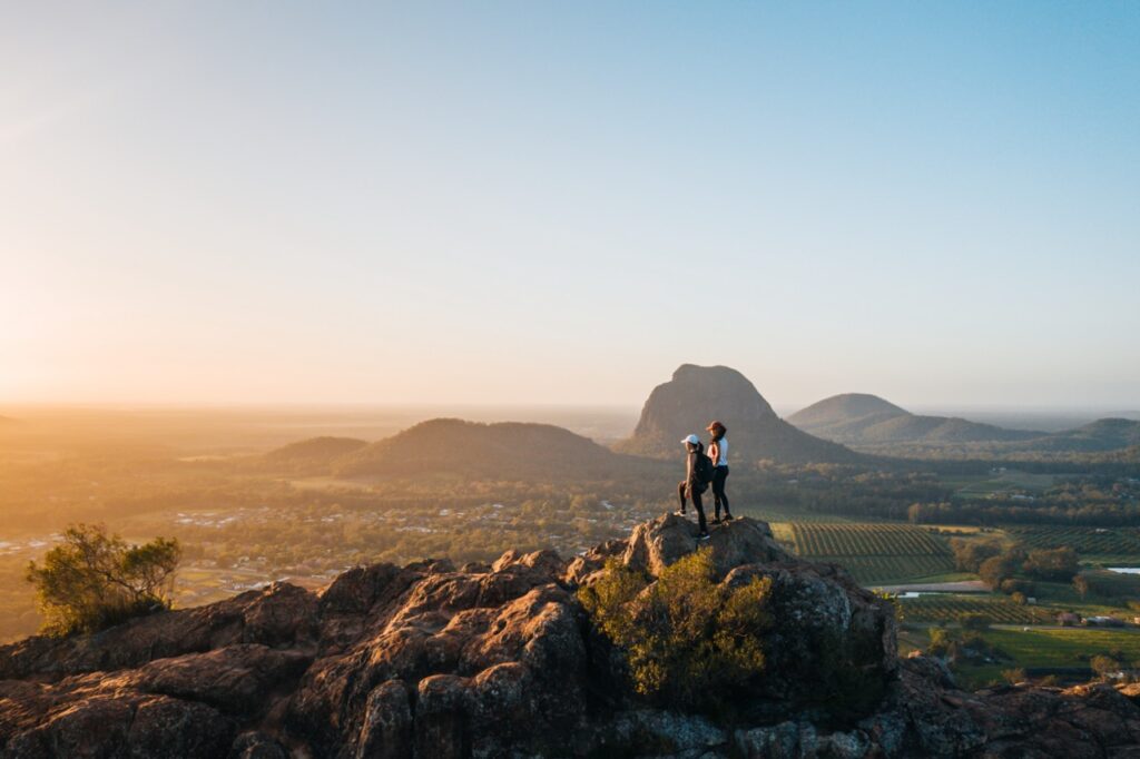 Two people stand on a rocky outcrop overlooking a landscape of fields and mountains during sunrise or sunset.