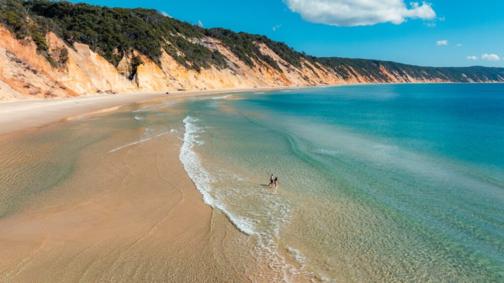 A couple walks along a deserted beach with eroded cliffs and clear blue water under a sunny sky.