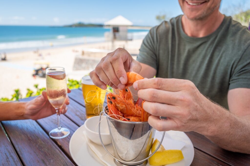 A person peels shrimp at an outdoor table near a beach, with a glass of champagne and a beer nearby.