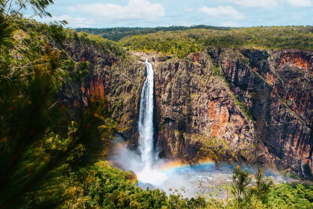 Tall waterfall cascading down a rocky cliff into a lush green forest, with a rainbow visible near the base.