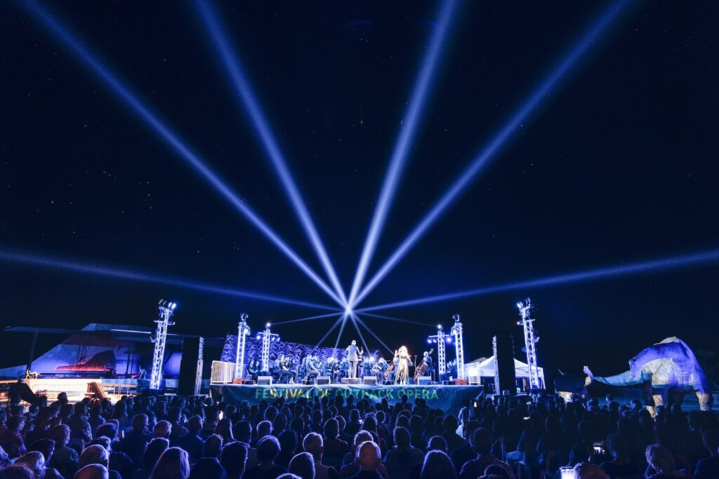 Outdoor concert stage with bright blue spotlights beaming into the night sky, audience seated in front, and festival decorations on stage.