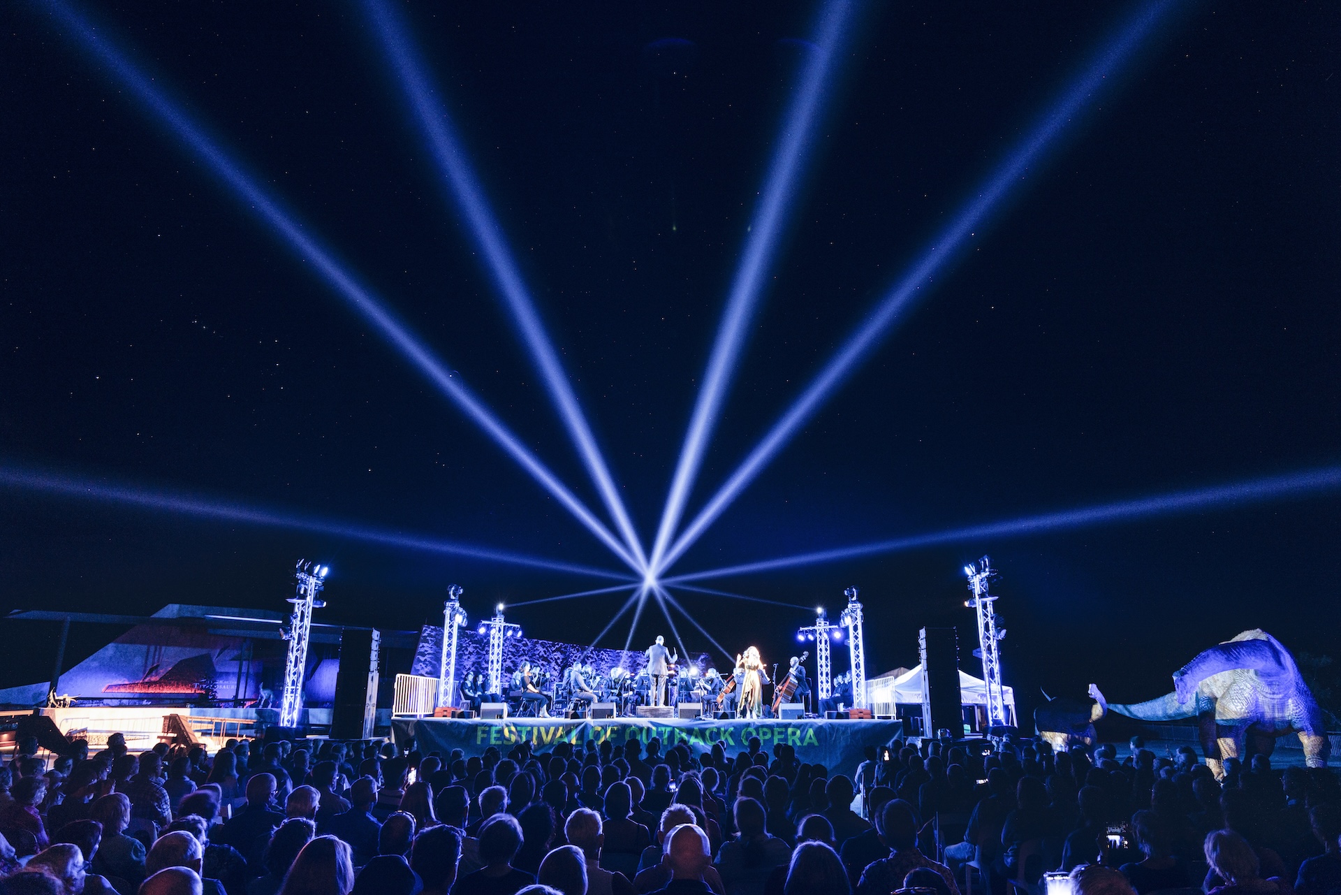 A large crowd watches a nighttime outdoor concert with blue spotlights radiating from the stage into the sky.