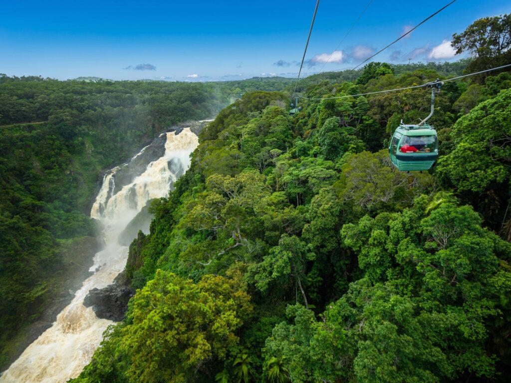 A cable car moves over dense rainforest toward a large waterfall, with lush green trees and blue sky in the background.