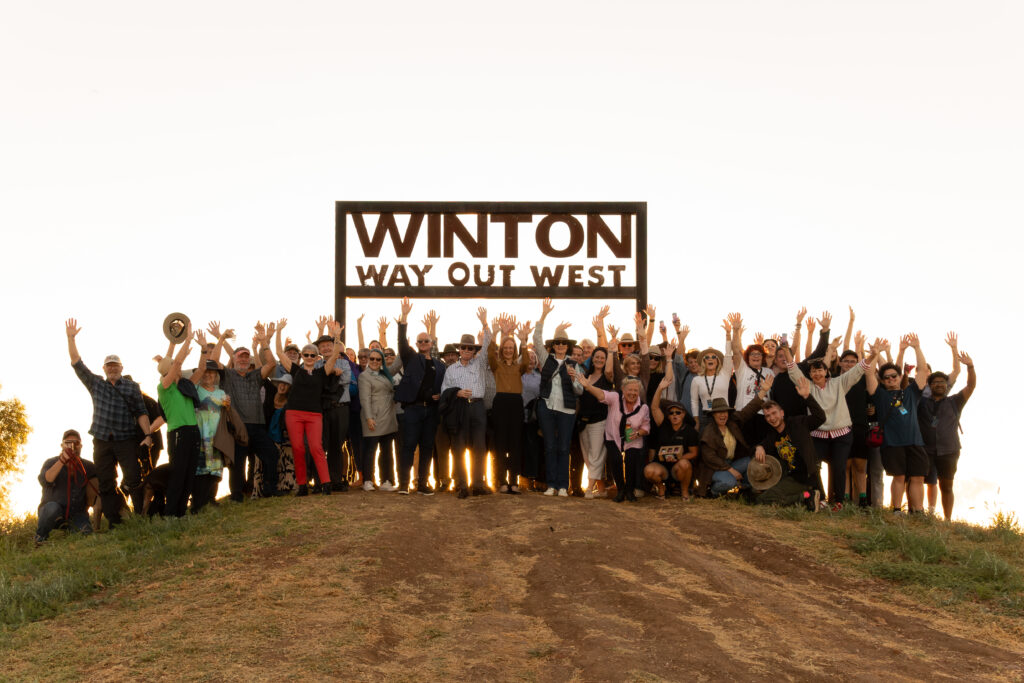 A large group of people stand on a hilltop in front of a sign reading 