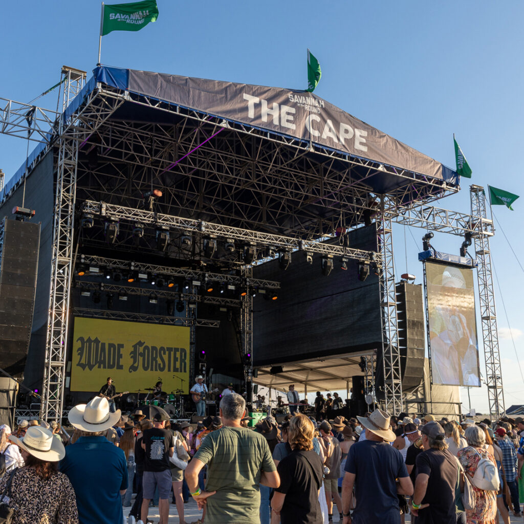 A crowd watches a band perform on an outdoor stage labeled 