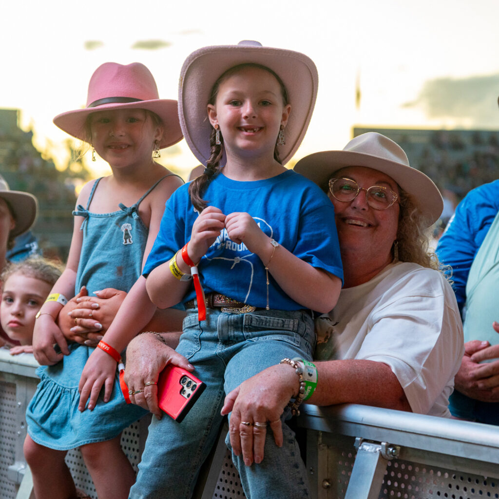 Three people wearing hats, including two children and one adult, stand and smile by a barrier at an outdoor event, as others look on in the background.