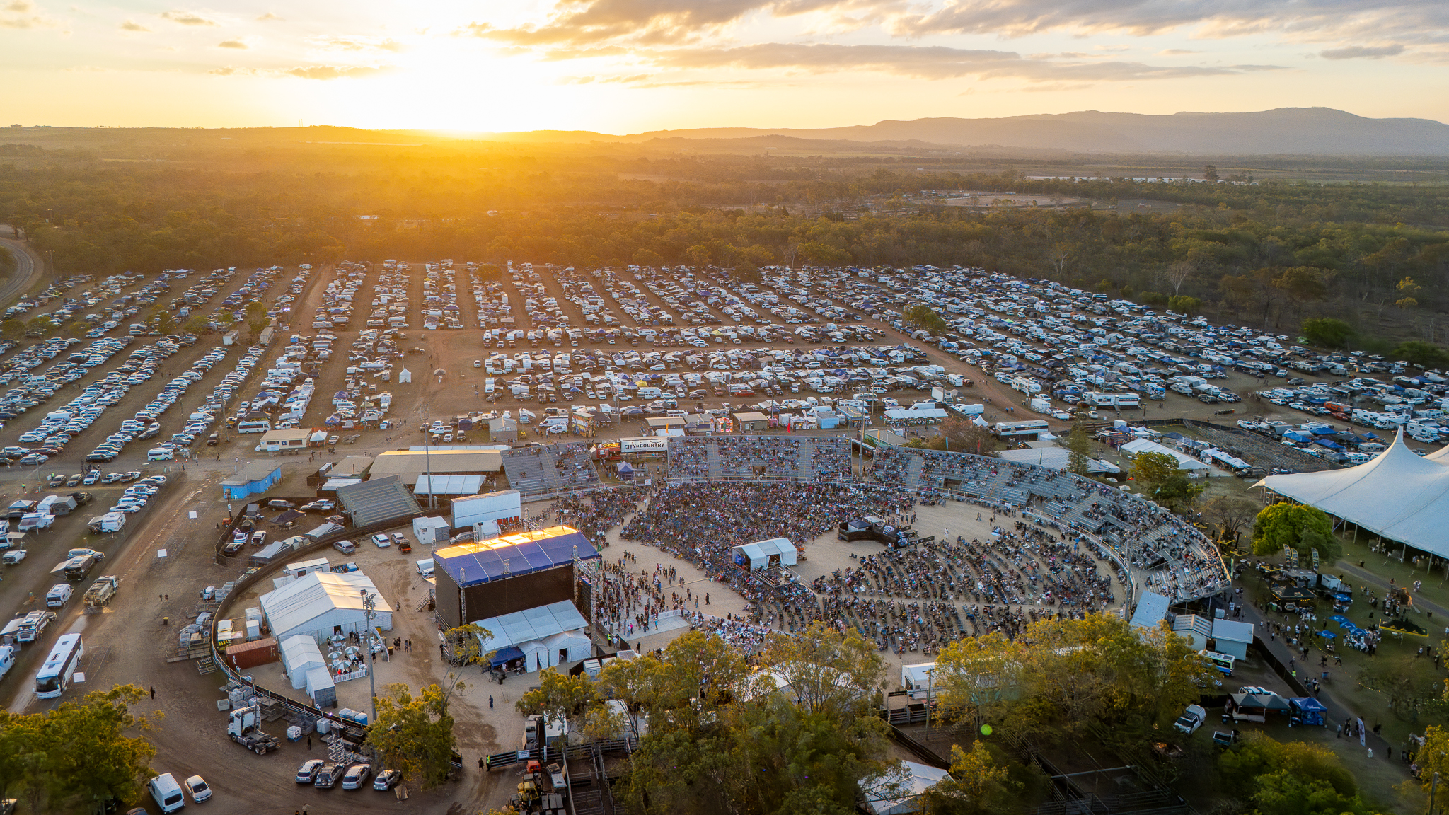 Aerial view of a large outdoor concert or event at sunset, with a full amphitheater and surrounding parking area filled with cars and RVs.