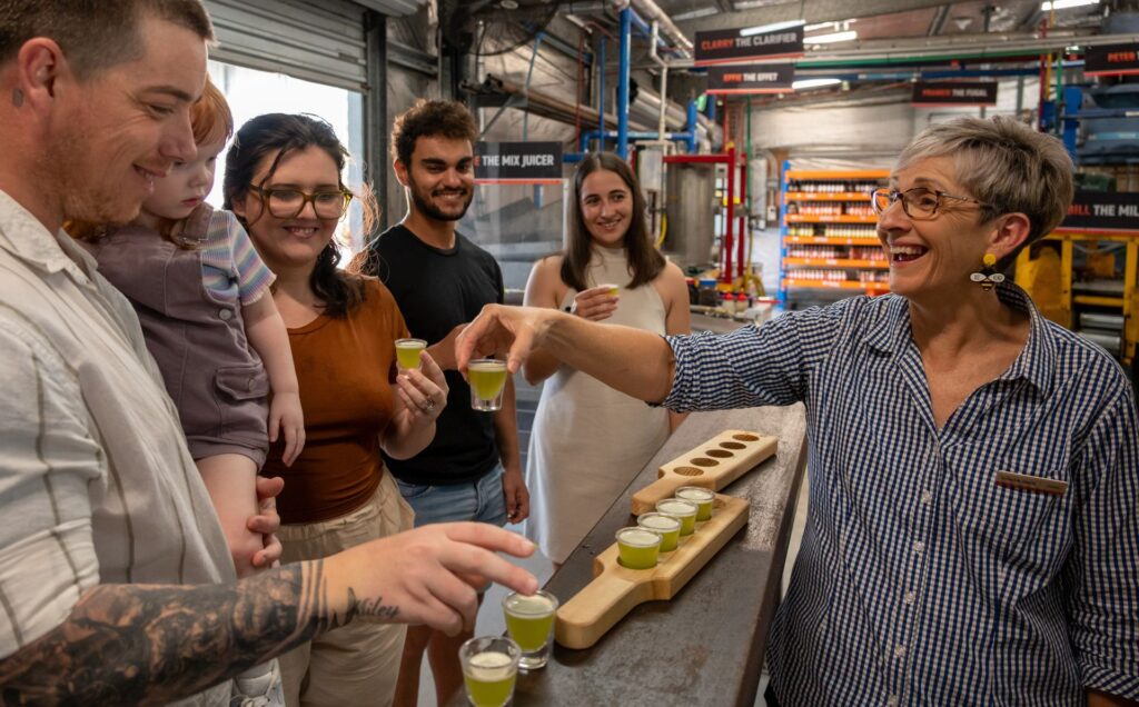 A group of people sample small glasses of green juice with a guide in an industrial facility, standing around a counter with a wooden tray of drinks.