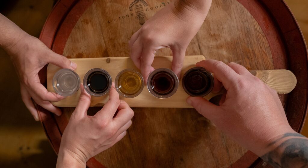 Three hands reach for five different colored drinks in small glasses arranged on a wooden paddle on top of a round wooden table.