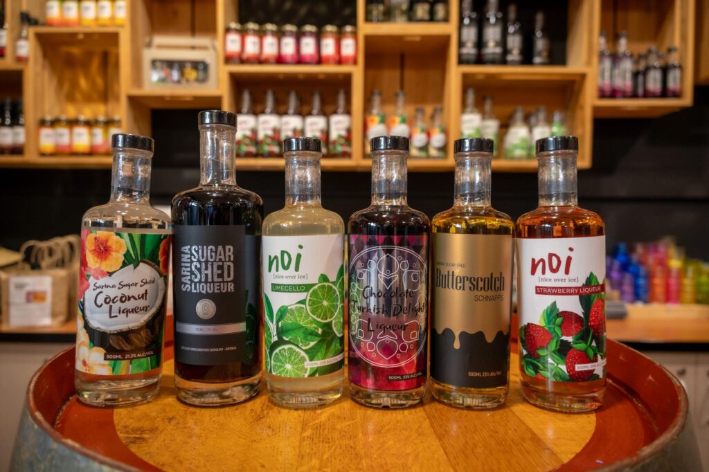 Six assorted bottles of liqueur are lined up on a wooden barrel with shelves of bottles and jars in the background.