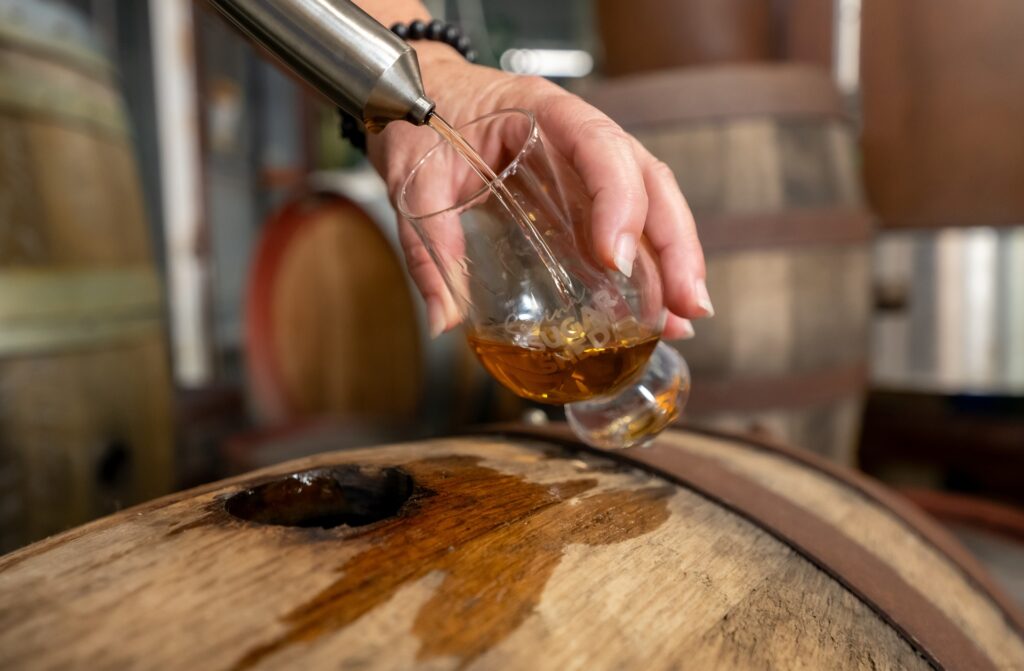 A hand holds a glass at an angle, filling it with whiskey from a barrel using a metal device, with wooden barrels visible in the background.