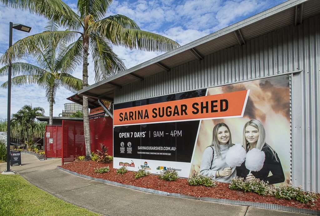 A large sign for Sarina Sugar Shed is displayed on a corrugated metal building, with two women holding cotton candy, palm trees, and a walkway visible nearby.