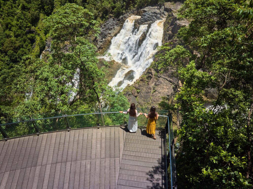 Two people stand on a wooden viewing platform overlooking a large waterfall surrounded by dense green forest.