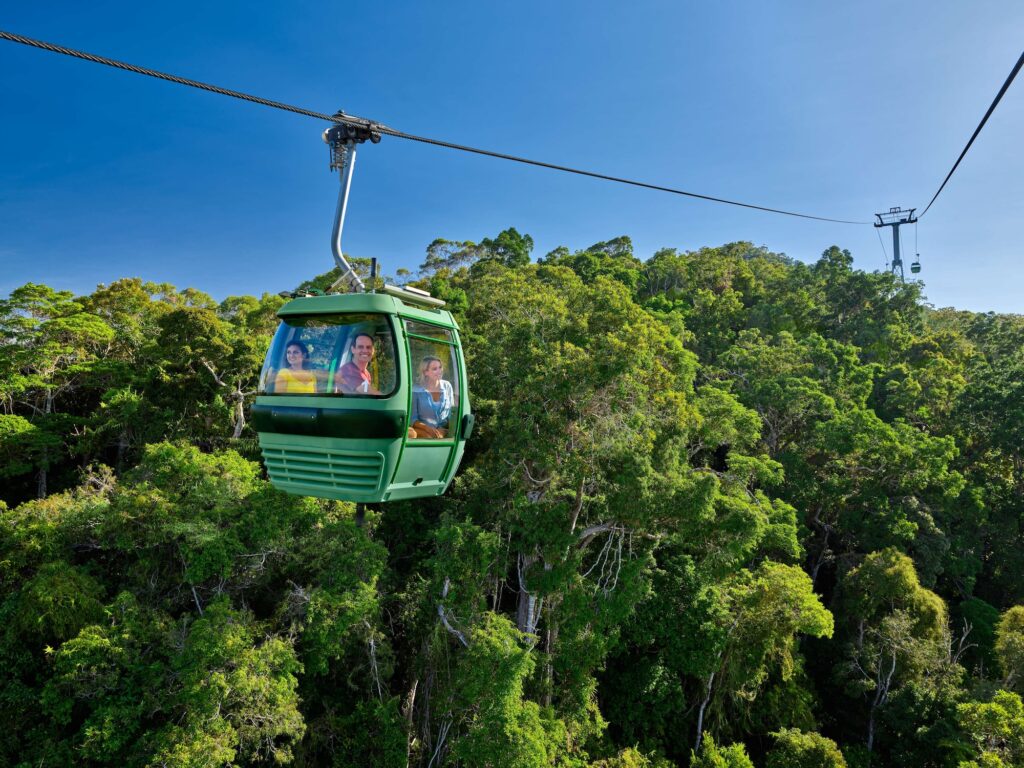 A green cable car travels above dense tropical forest with three people inside, under a clear blue sky.