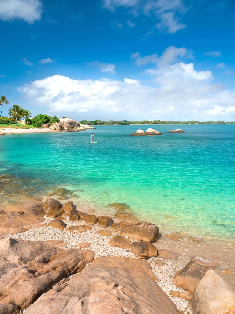 A person stands on a paddleboard in clear, turquoise waters near a rocky shore with scattered vegetation. The sky is partly cloudy.