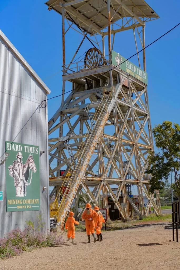 Three people in orange protective clothing walk past a tall mining structure labeled 