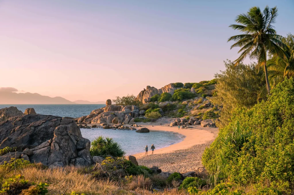 A secluded beach with two people walking along the shore, surrounded by rocky terrain, palm trees, and vegetation, under a clear sky at sunset.