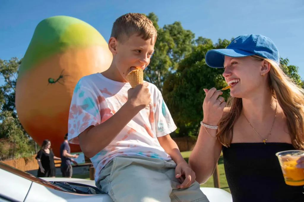 A boy sits on a car eating an ice cream cone while a girl in a blue cap enjoys a treat next to him. In the background, there is a large mango sculpture and people walking by.